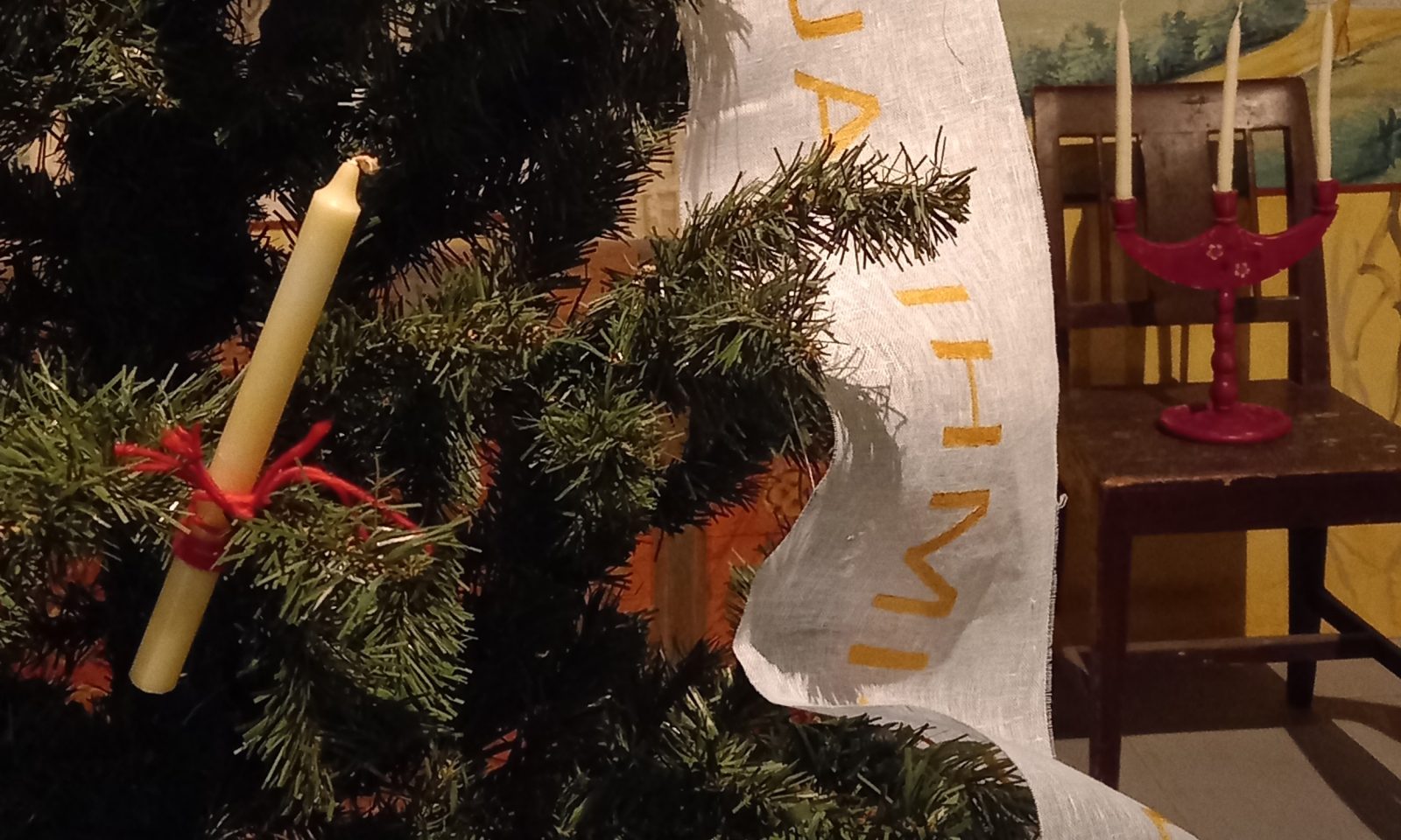 Lähikuvassa joulukuusen oksa, johon sidottu punaisella nauhalla ohut ja pitkä kynttilä. Joulukuusessa myös valkoinen leveä tekstitetty nauha. Nauhasta näkyy vain muutamia kultaisia kirjaimia. Taustalla ruskean tuolin päälle asetettu punainen kynttelikkö, jossa kolme pitkää valkoista kynttilää.