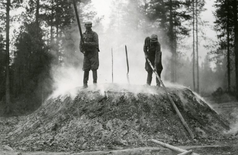 Kaksi miestä piiskaamassa savuavaa tervahautaa.