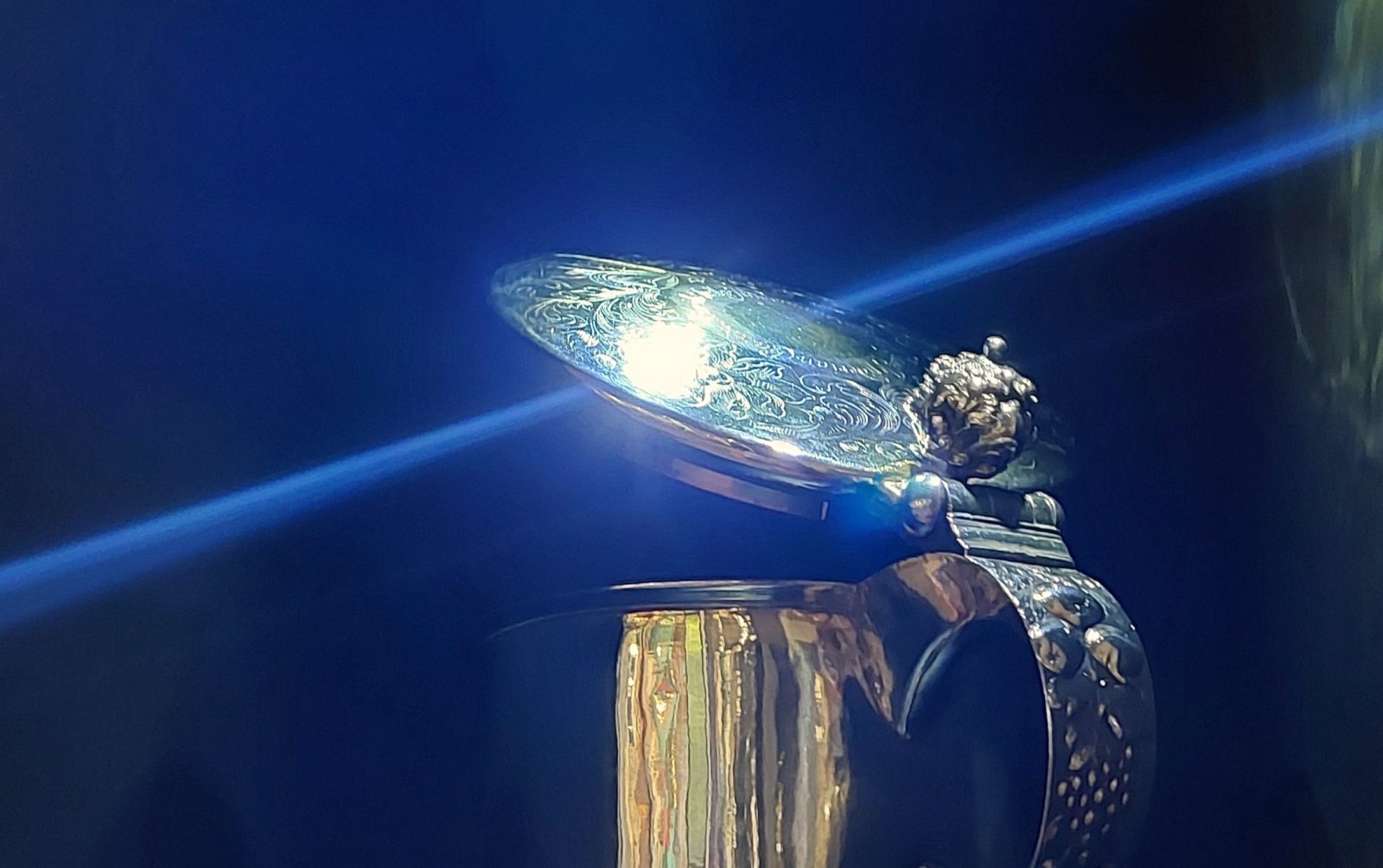 Tummalla taustalla hopeinen kannu, jonka kannesta valon säteet heijastuu sivuille.