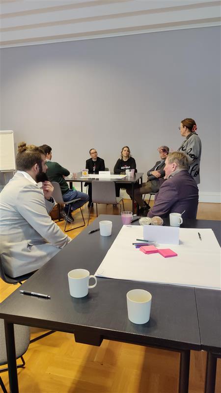 Ihmisiä istuu pöytien ääressä ja yksi puhuu. Pöydällä kahvikuppeja ja iso valkoinen paperi, johon on liimattu post it -lappuja.