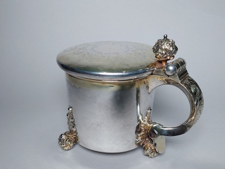 Hopeinen kannu, jossa kansi, iso kahva ja koristeelliset jalat.