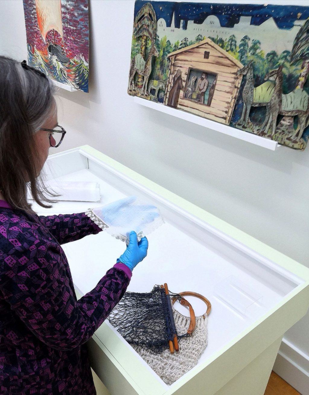 Konservaattori asettelee tekstiilejä näyttelypöydälle. Pöydän yläpuolella tauluja. Kuva otettu Oulun taidemuseolla.