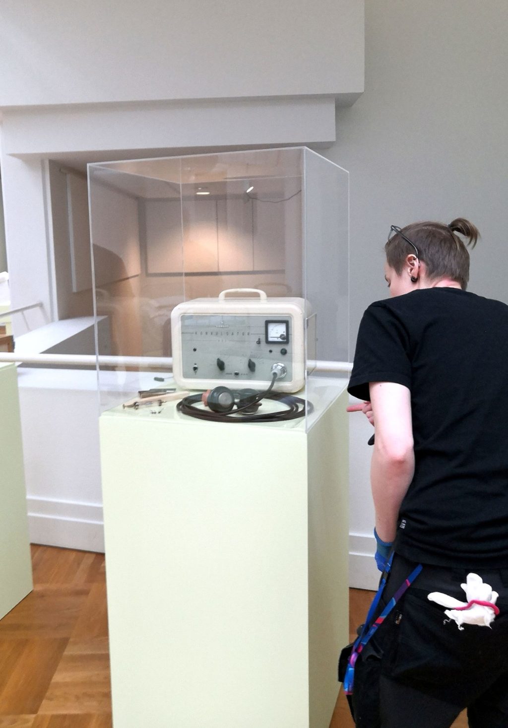 Museon henkilökuntaan kuuluva ihminen tarkastelee lasivitriinissä olevaa valkoista sähköshokkilaitetta. Kuva otettu Oulun taidemuseolla.