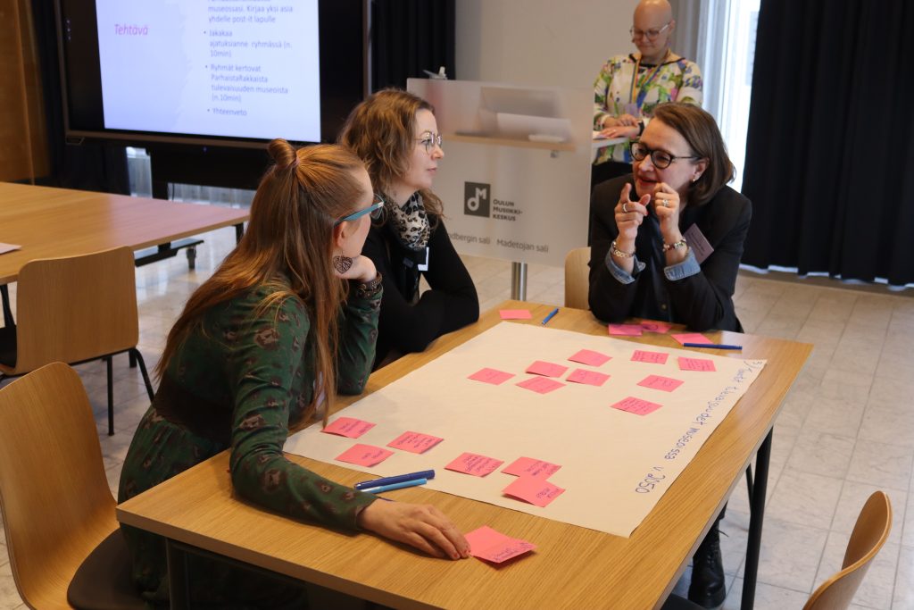 Kolme naista pöydän ääressä juttelevat. Pöydällä iso valkoinen paperi, johon liimattuna pinkkejä post it -lappuja ja kirjoitusta.