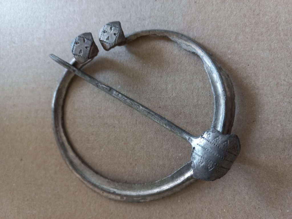 Pyöreä hopeinen solki, keskellä halkoo neula.