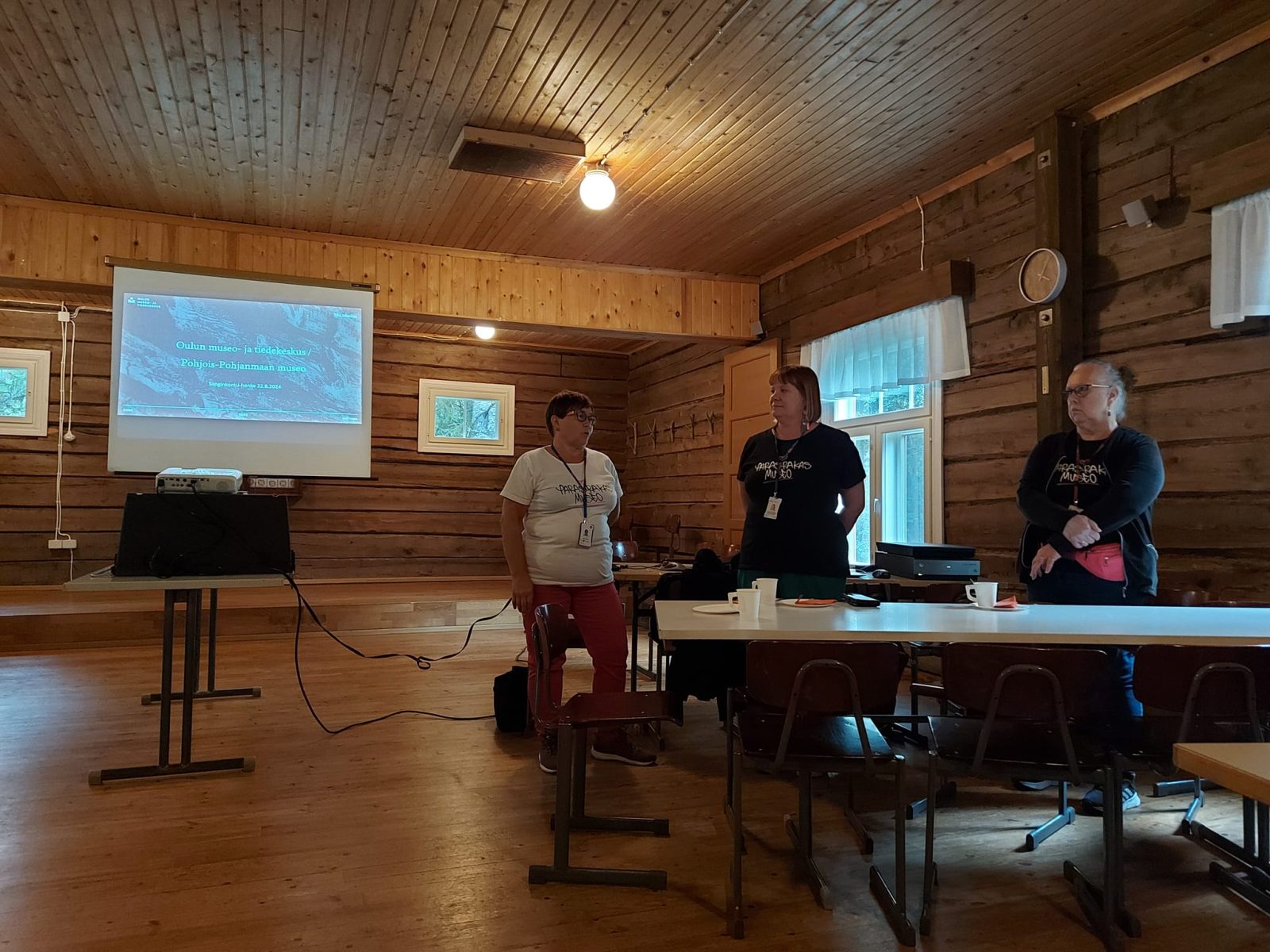 Hirsirakennus sisältä. Kolme naista seisoo rivissä, taustalla valkokankaalla esitys. Naisilla paidat päällä, jossa lukee: ParasRakas museo.