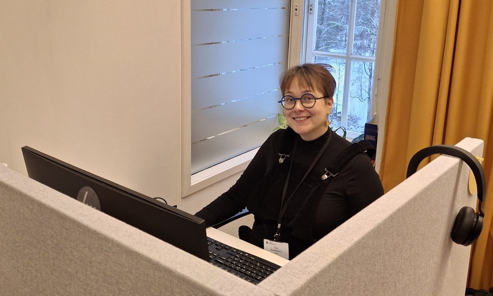 Nainen lyhyissä ruskeissa hiuksissa ja silmälaseissa istuu tietokoneen edessä ja hymyilee.