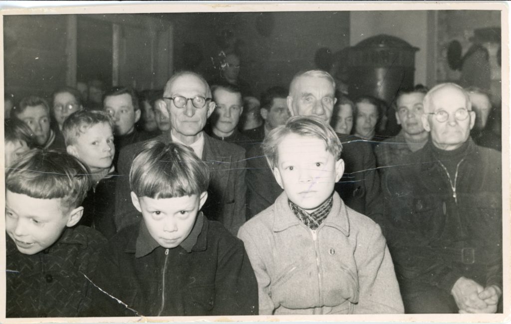 Vanha mustavalkoinen kuva. Ihmisiä istumassa kuvaajaa kohti. Poikia eturivissä, miehiä heidän takanaan.