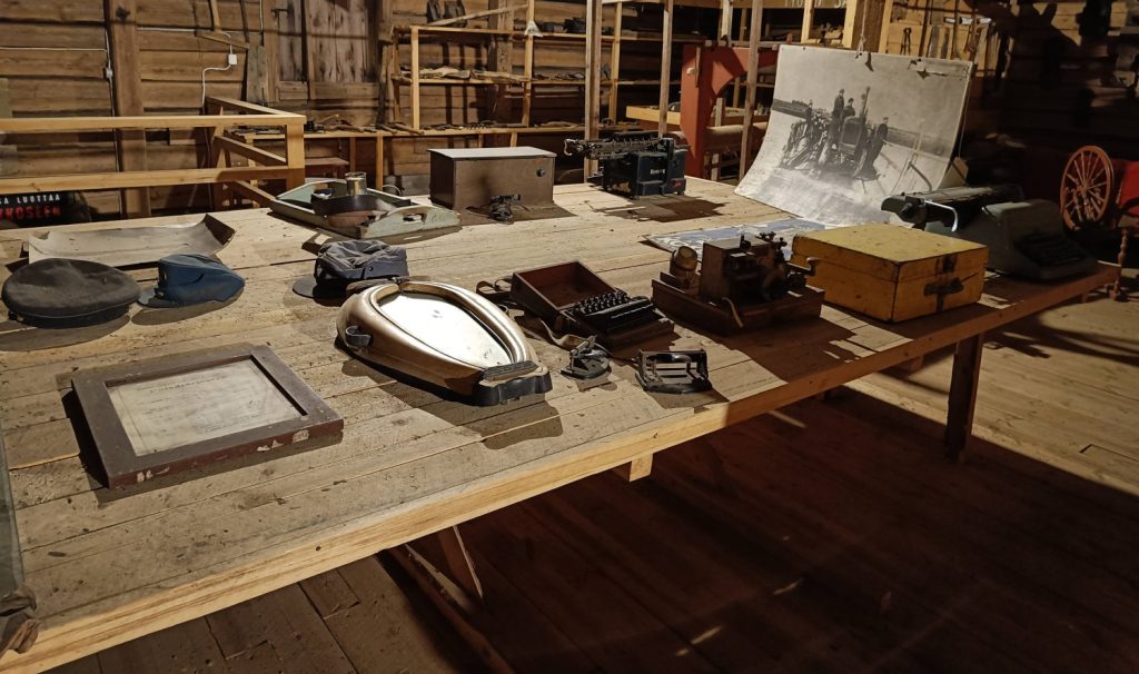 Puisen rakennuksen sisällä puinen pöytä, jonka päällä erilaisia vanhoja tavaroita. Esim. lakkeja, peili.