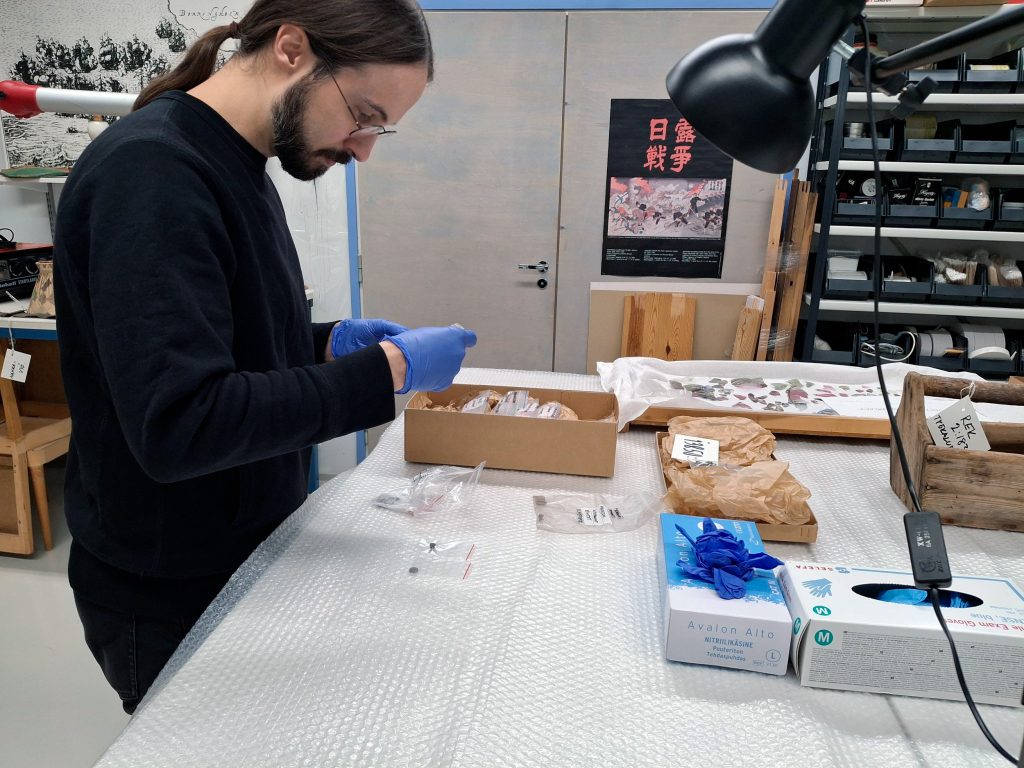 Mies tutkii jotain käsissään olevaa. Kädessä hänellä siniset kumihanskat ja pöydällä muovin päällä tavaroita pienissä muovipusseissa.