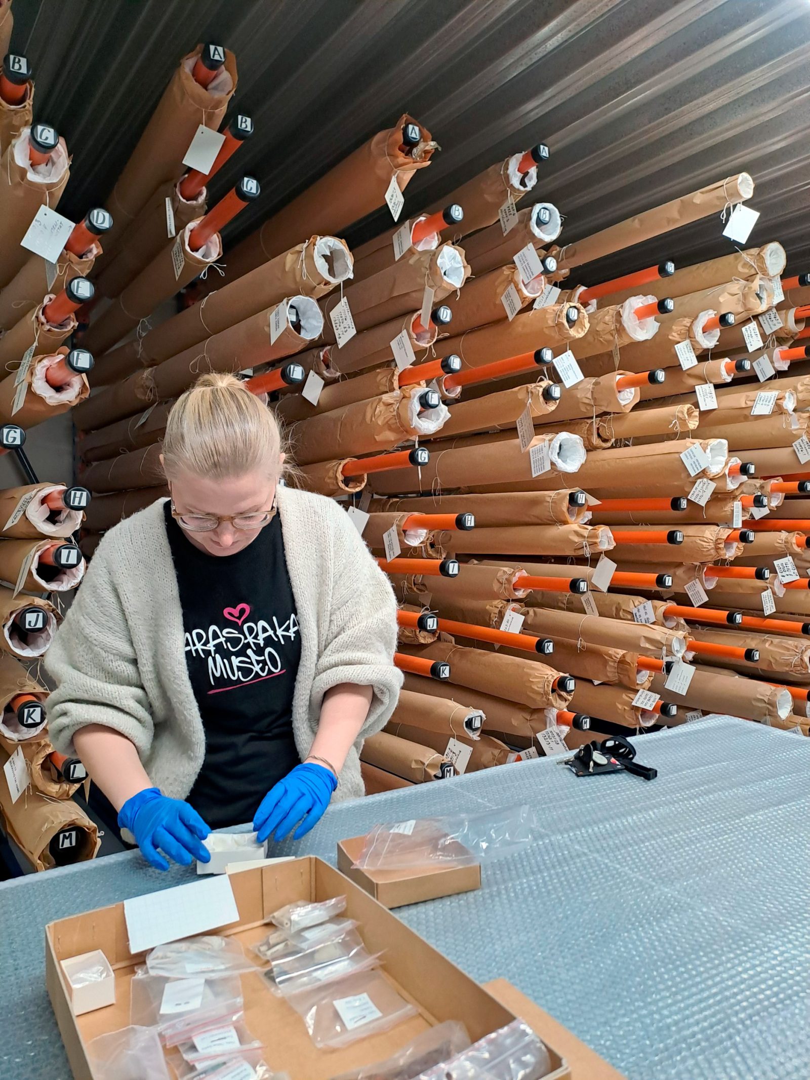 Nainen tutkii jotain käsissään sinisillä kumihanskoilla. Edessä pöydällä muovin päällä pahvilaatikko, jossa pussitettuja pieniä esineitä.