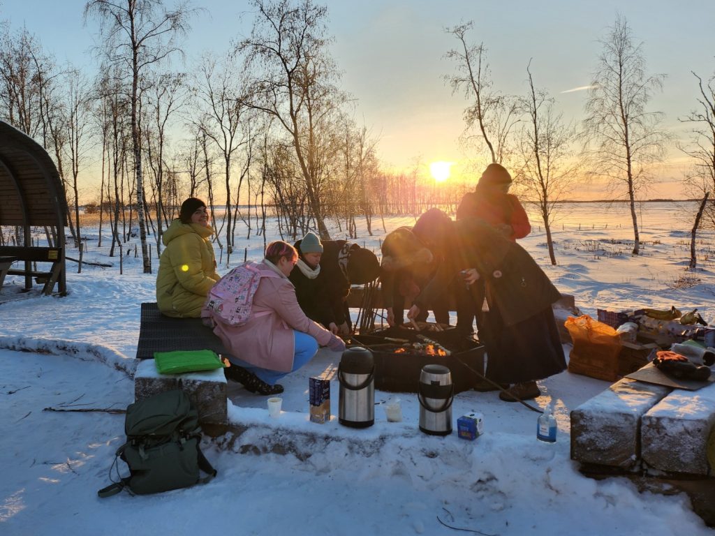 Ihmisiä nuotiolla, auringonlasku horisontissa. Mukana ihmisillä on eväitä ja retkitarvikkeita.