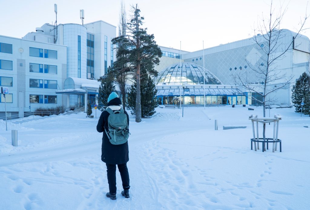 Valkoinen iso tiilinen rakennus. Lunta maassa, ihminen seisoo selin kameraan, katse kohti rakennusta.