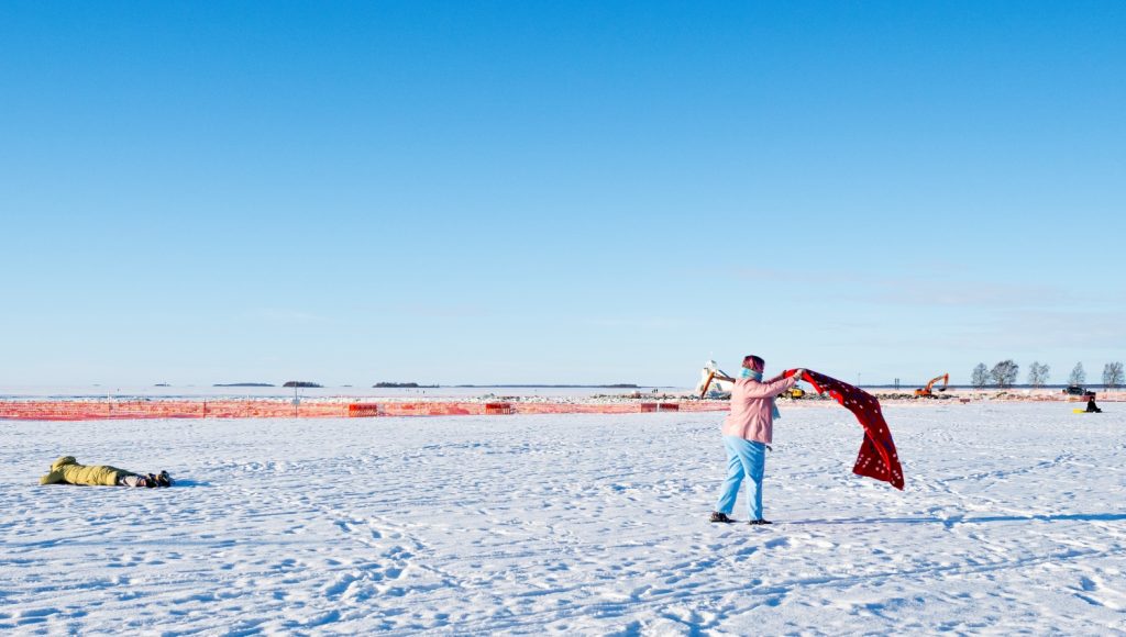 Ihminen vaaleanpunaisessa takissa ja vaaleansinisissä housuissa levittää huopaa lumiselle rannalle. Taustalla työmaa-aitoja ja kaivinkoneita sekä horisontissa meri ja saaristoa.