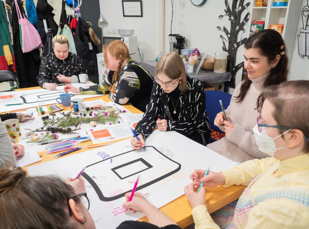 Ihmisiä pöydän äärellä. He kirjoittavat pöydällä olevaan isoon paperiin omilla kynillään.