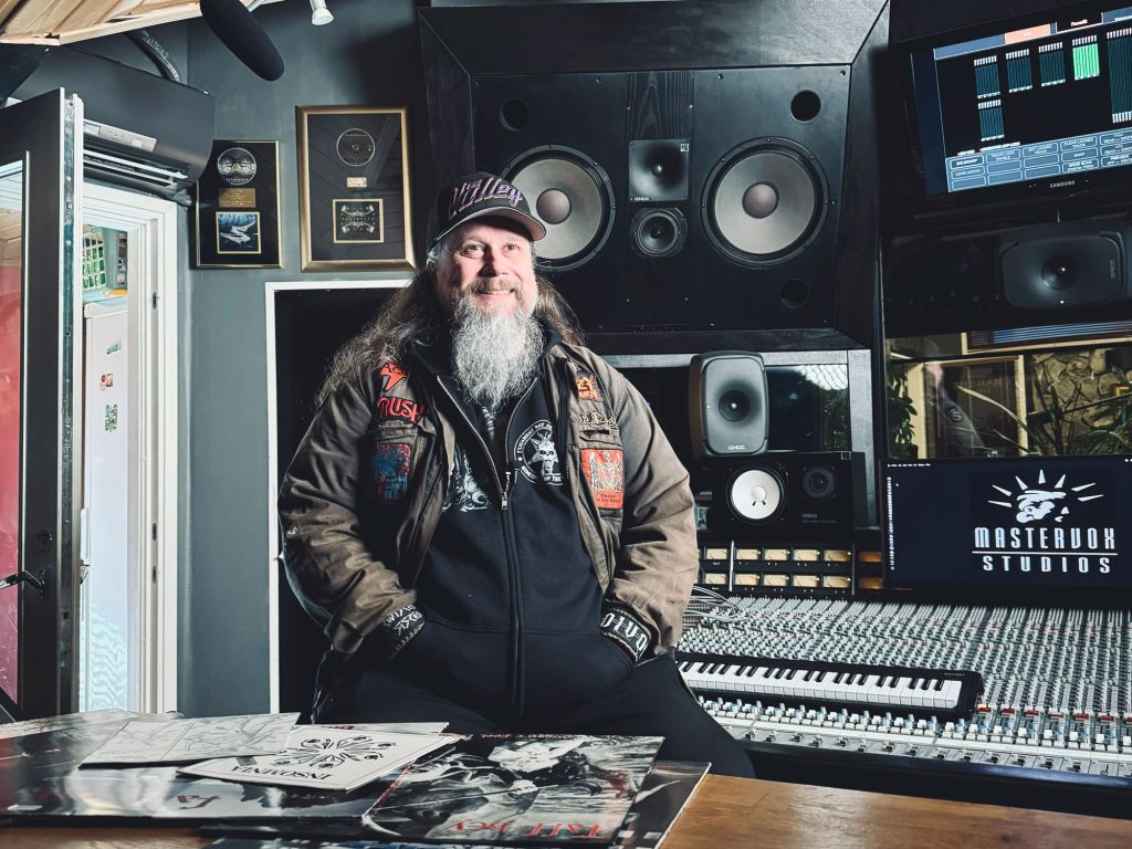 Henkilö mustassa hupparissa ja ruskeassa takissa, jossa on paljon bändimerkkejä istumassa miksauspöydän edessä.