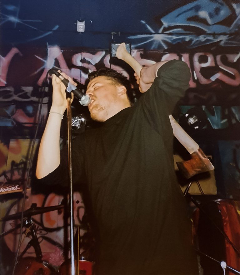 Mikko Laurila laulaa ja pitelee kirvestä selkänsä takana. Taustalla keikkapaikan graffitein koristeltua tummaa seinää.