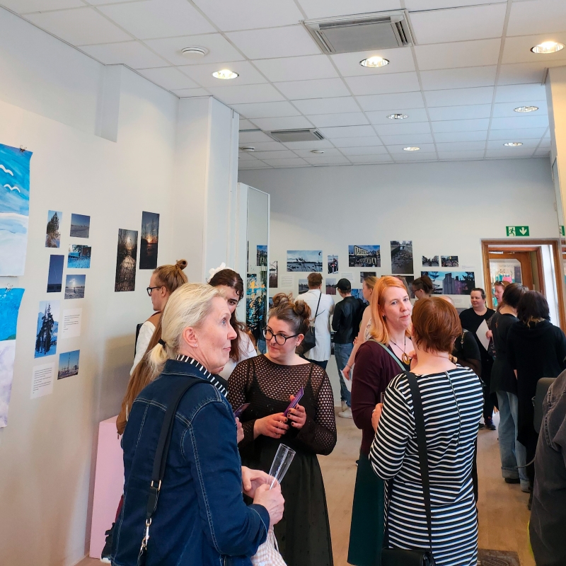 Ihmisiä näyttelytilassa. Seinillä valokuvia ja muita taideteoksia.