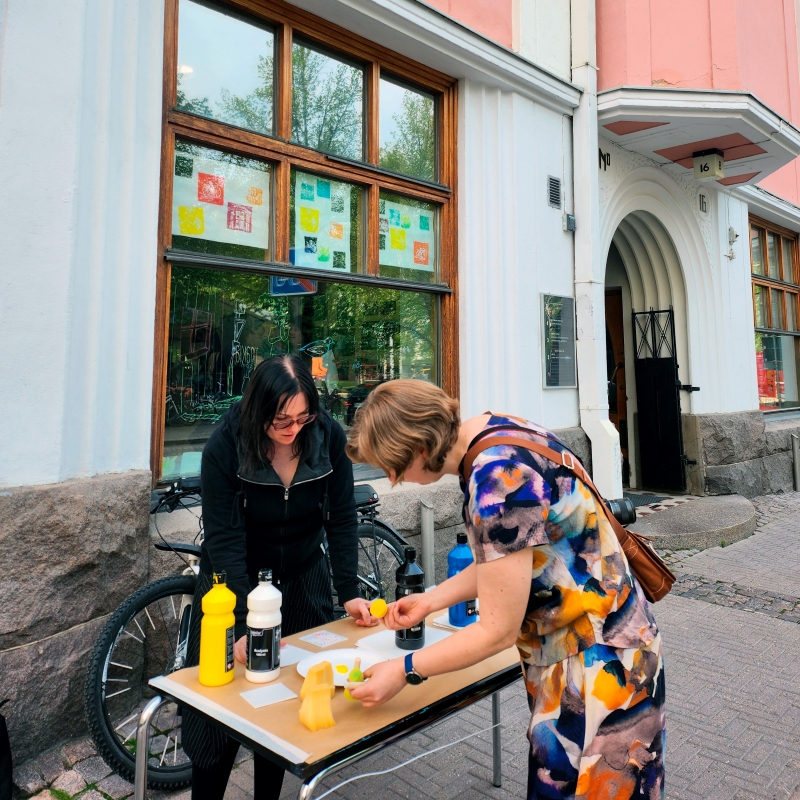 Kivisen rakennuksen edessä pihalla pöytä, jossa askartelutarvikkeita. Kaksi ihmistä kumartuneena pöydän ääreen.