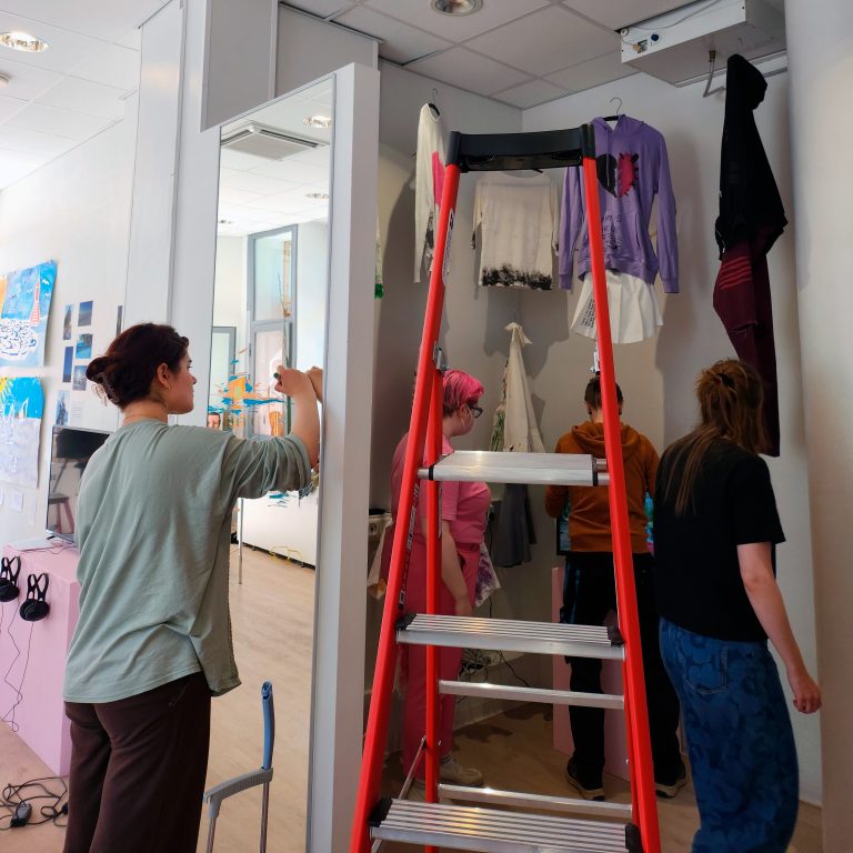 Punaiset tikkaat ja niiden takana paitoja ripustettu kattoon henkareilla. Neljä ihmistä tikkaiden ympärillä.