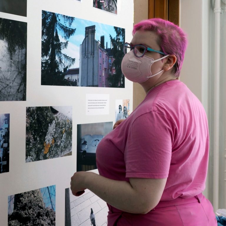 Pinkkipaitainen nainen, lyhyet pinkit hiukset ja hengityssuojain kasvoillaan. Seinällä valokuvia.