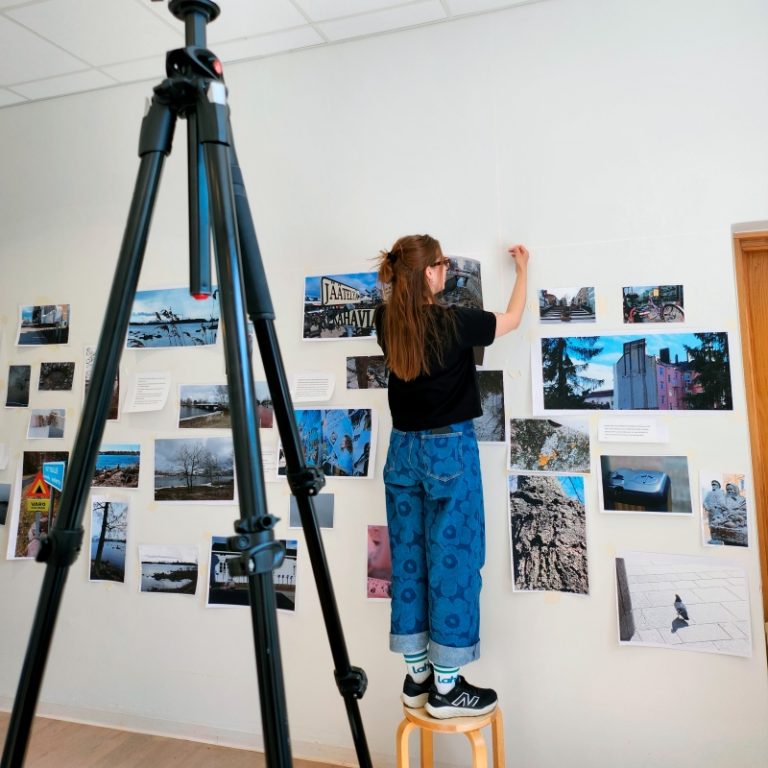 Nainen seisoo tuolilla ja kiinnittää seinälle lisää valokuvia. Kuvan edustalla kameran jalusta.