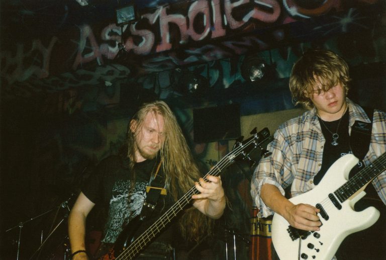 Kitaristi Miika Tenkula soittaa kuvan oikeassa laidassa ja basisti Niko Karppinen keskellä kuvaa. Taustalla keikkapaikan tummaa graffitein koristeltua ympäristöä.