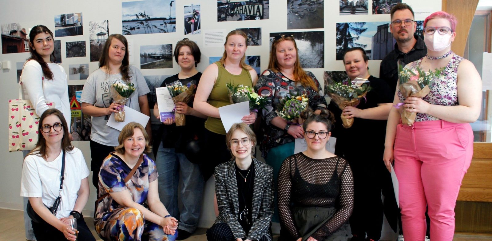 Ihmisiä kukat kädessä ryhmittyneenä yhteiskuvaan. Taustan seinällä valokuvia.