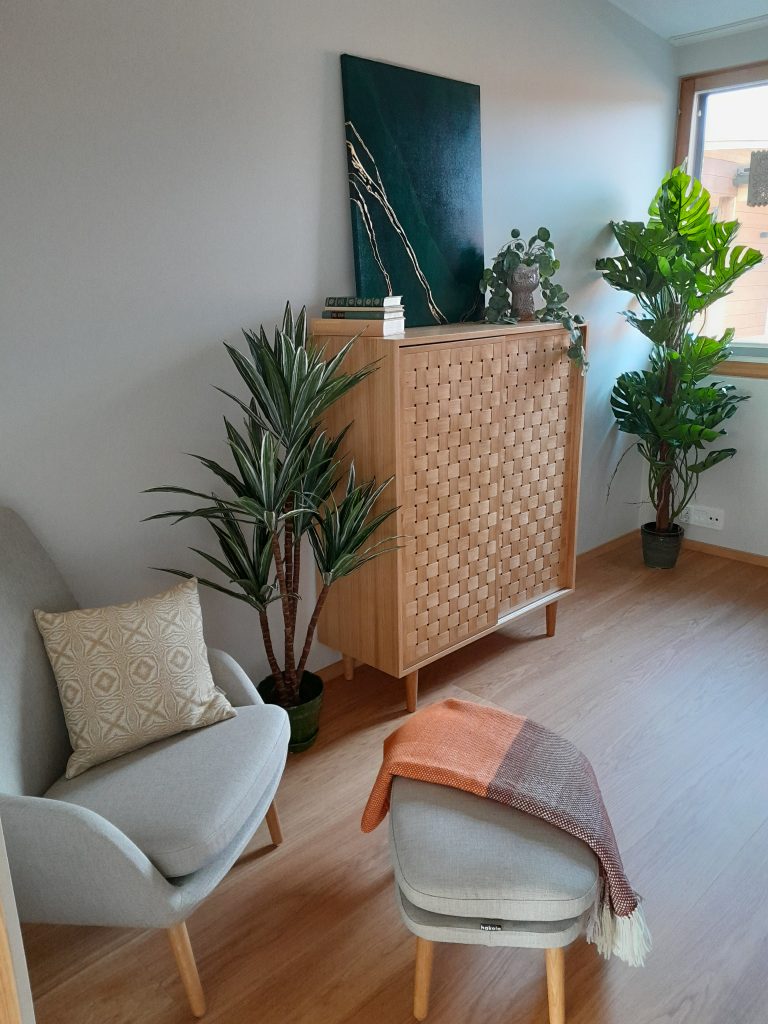 Huone, jossa nojatuoli ja senkki, sisustus vaaleaa puuta ja vaaleita sävyjä tekstiileissä, viherkasveja paljon.