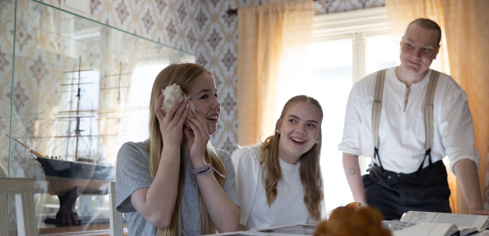 Kolme henkilöä pöydän ympärillä, yksi kuuntelee isoa simpukkaa.