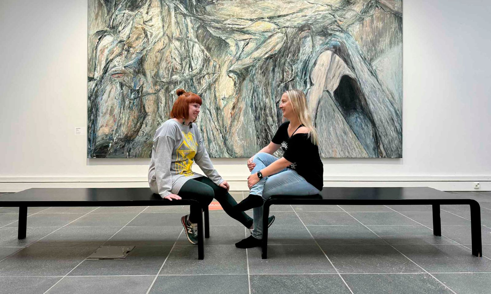 Kaksi henkilöä istuu penkeillä kasvotusten, takana taideteos.