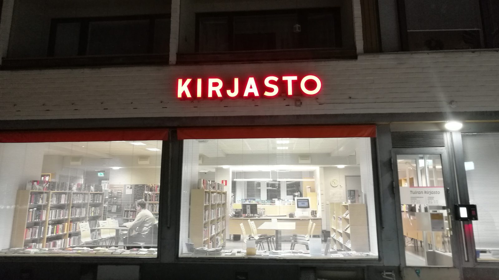Ikkunasta näkyy kirjasto. Ikkunan päällä punaisilla ja valaistuilla kirjaimilla lukee KIRJASTO.