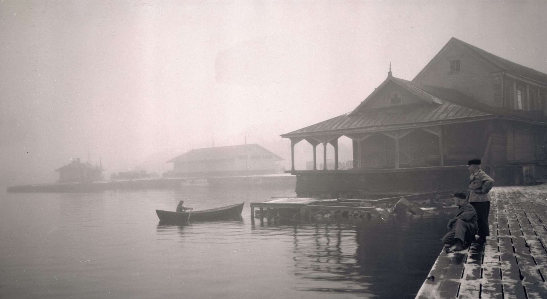 Mustavalkoisessa, vanhassa kuvassa kaksi poikaa seisoo laiturilla. Taustalla vanha puinen rakennus ja soutuvene on vedessä.