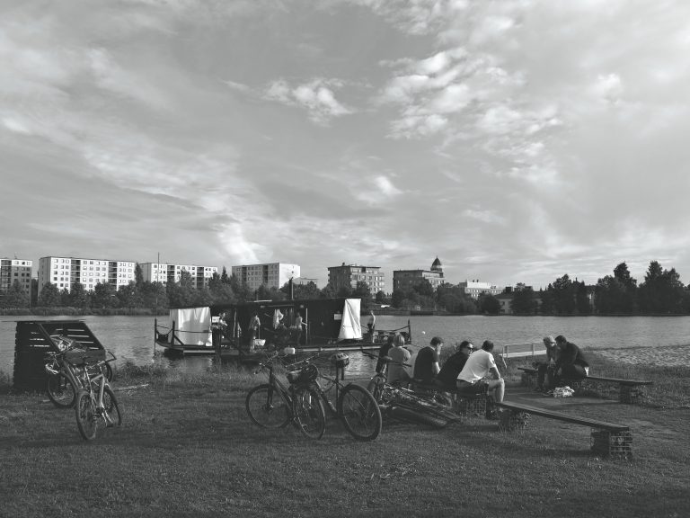 Henkilöitä joen rannalla, vieressä kelluu saunalautta, jossa saunojia.