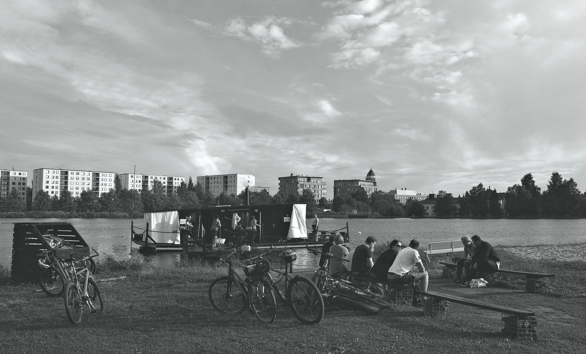 Saunalautta Oulujoella Tuiran uimarannalla. Etualalla ihmisiä ja polkupyöriä. Taustalla kaupunkimaisemaa ja Oulun museo- ja tiedekeskuksen torni.