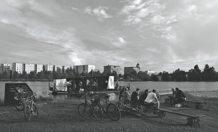 Saunalautta Oulujoella Tuiran uimarannalla. Etualalla ihmisiä ja polkupyöriä. Taustalla kaupunkimaisemaa ja Oulun museo- ja tiedekeskuksen torni.