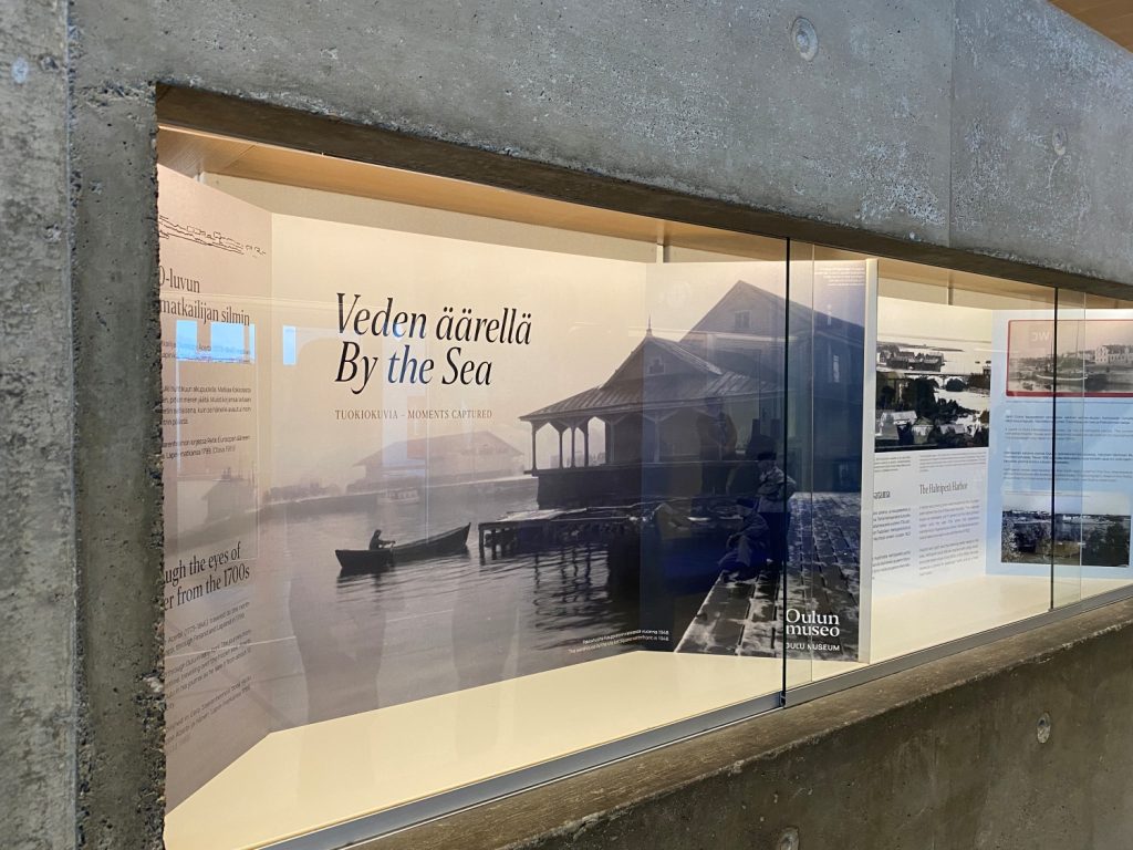 Betonisessa vitriinissä iso kuva, jossa teksti: Veden äärellä. By the sea.