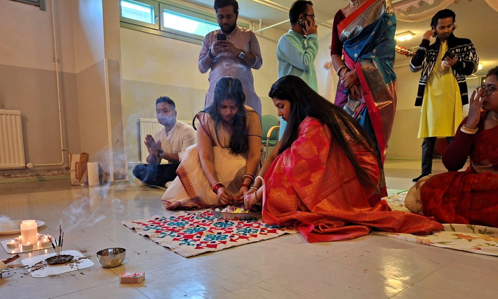 Kaksi naista intialaisisa juhla-asuissa kyykistyneenä lautasen ääreen. Taustalla ihmisiä intialaisissaj juhla-asuissaan.