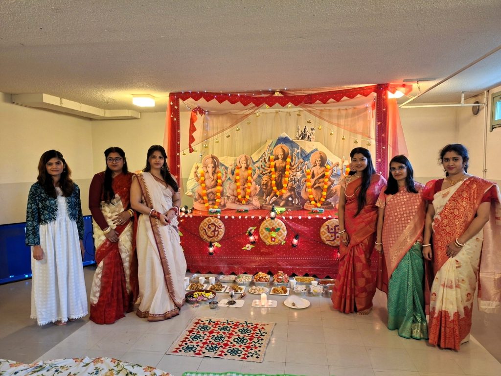 Naisia intialaisissa juhla-asuissaan asettuneena seisomaan alttarin molemmin puolin. Punaisella verhoilla kehystetyn alttarin sisällä on intialaisia kuvia koristeltuna. Lattialla edessä on erilaisia lautasia.