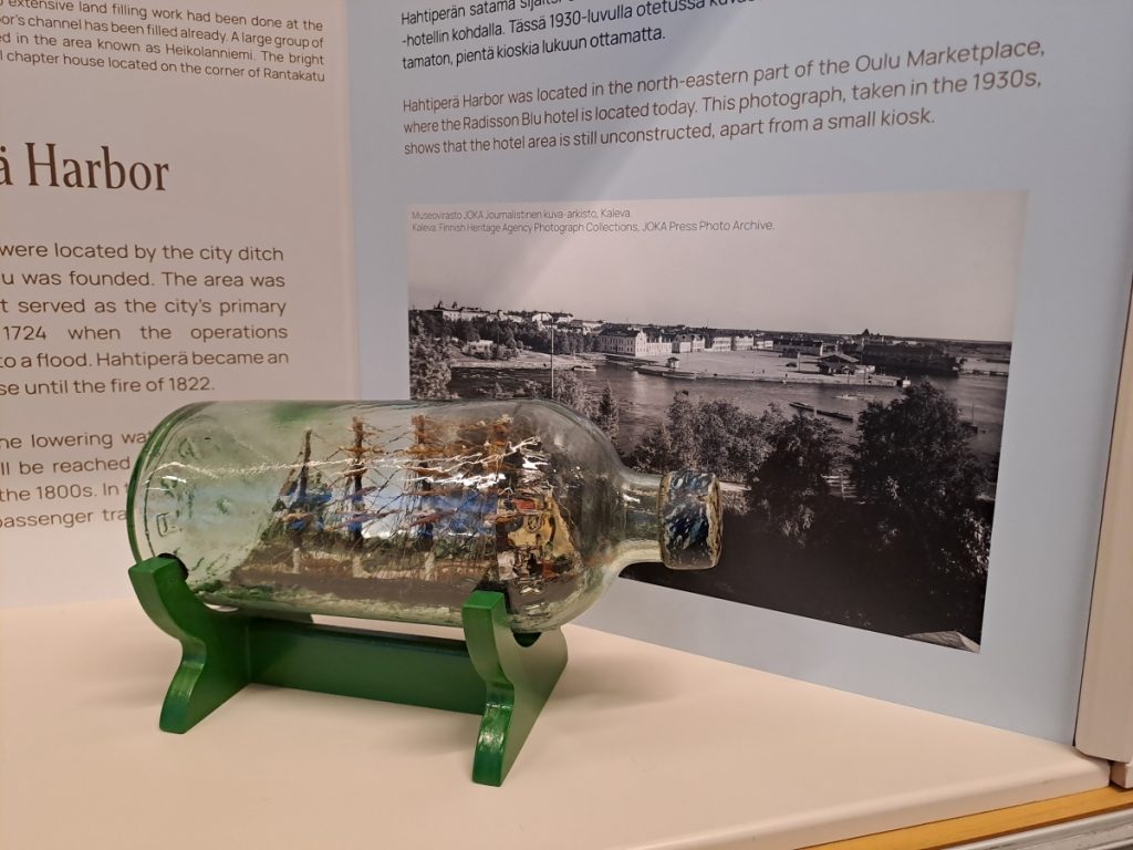 Vitriinissä vanha lasipullo telineessä. Pullon sisällä veneen pienoismalli.