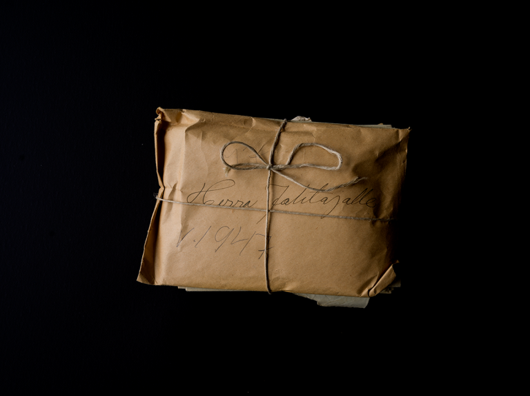 Ruskeaan paperiin narulla sidottu kirjenippu tummalla taustalla. Päällimmäiseem kuoreen raapustettu kaunokirjoituksella "Herra johtaja 1947".