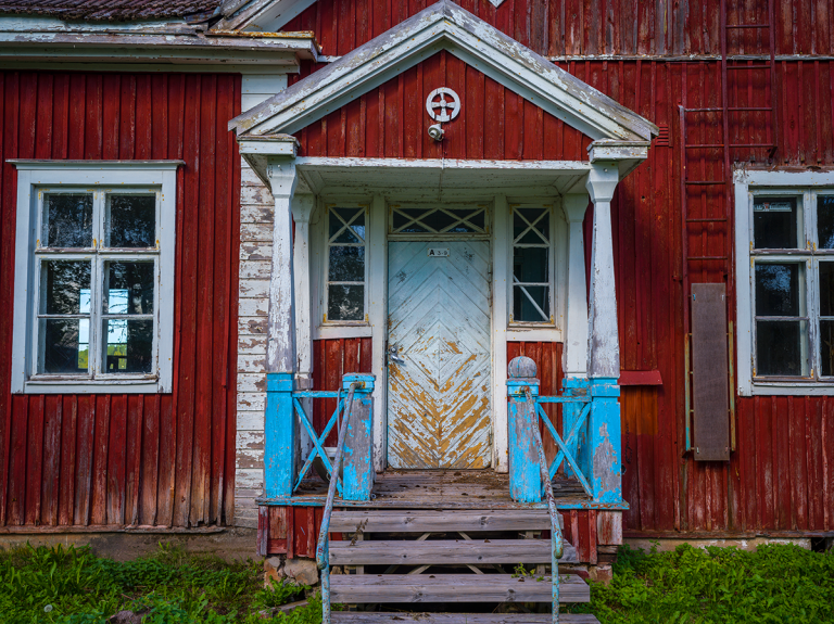 Vanhan punaisen puutalon sisäänkäynti. Kulunut valkoinen ovi, siniseksi maalatut kaiteet ja rapistunut kuisti.