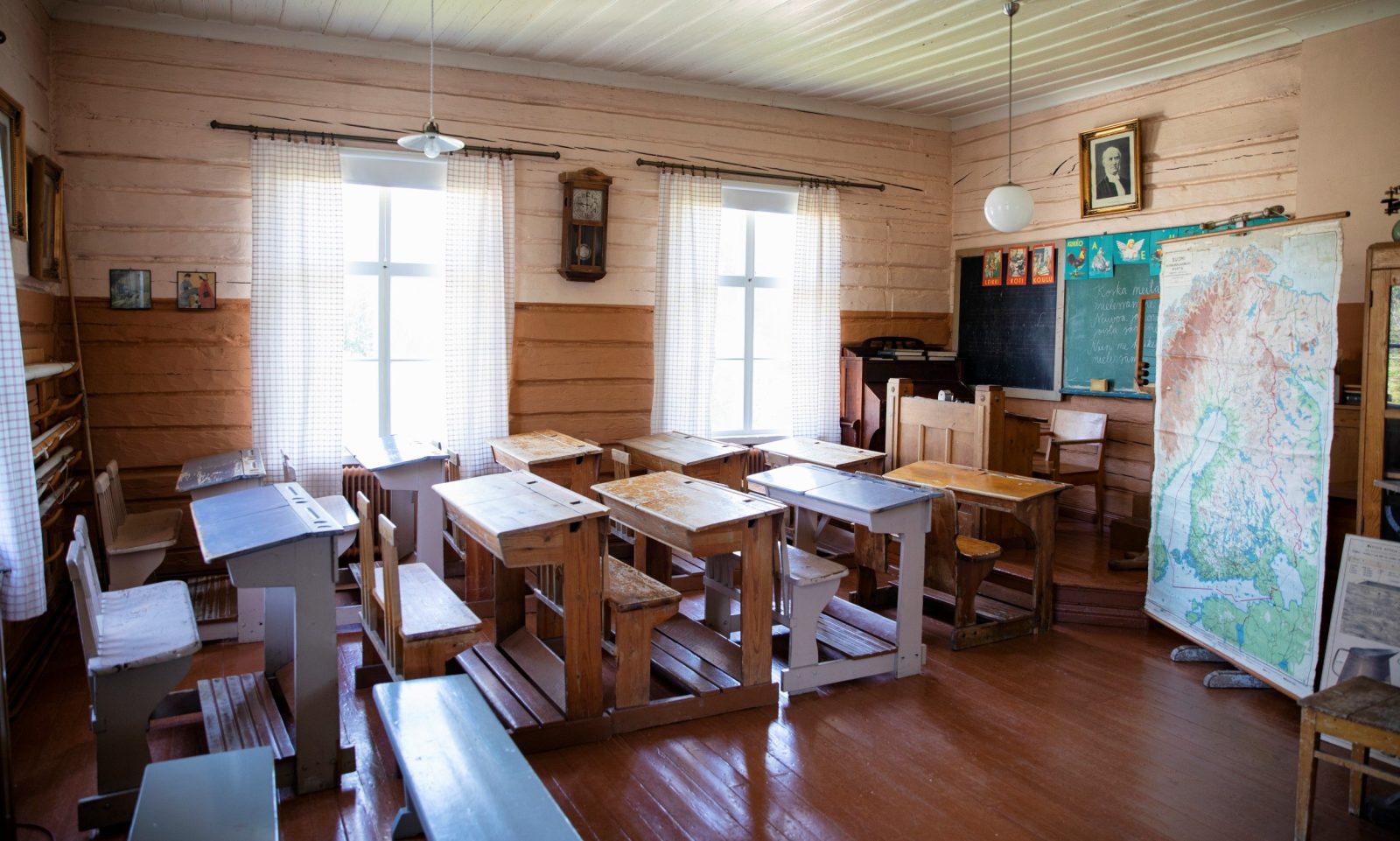 Vanha luokkahuone, jossa puisia pulpetteja, vihreä liitutaulu sekä telineessä roikkuva Suomen kartta.