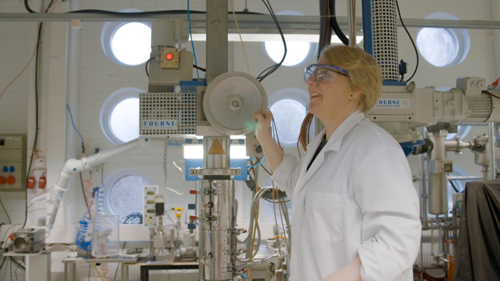 Valkoiseen takkiin ja suojalaseihin pukeutunut henkilö laboratoriossa.