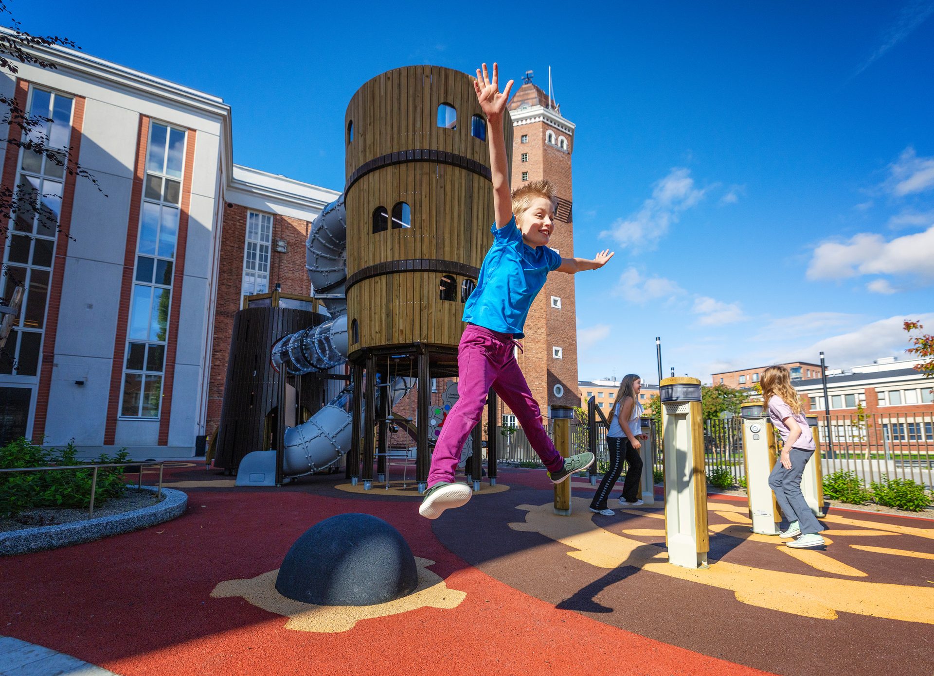 A child jumps on a balancing element at a playground in front of a large climbing structure resembling a castle tower. In the background, two other children and red brick buildings are visible on a sunny day.