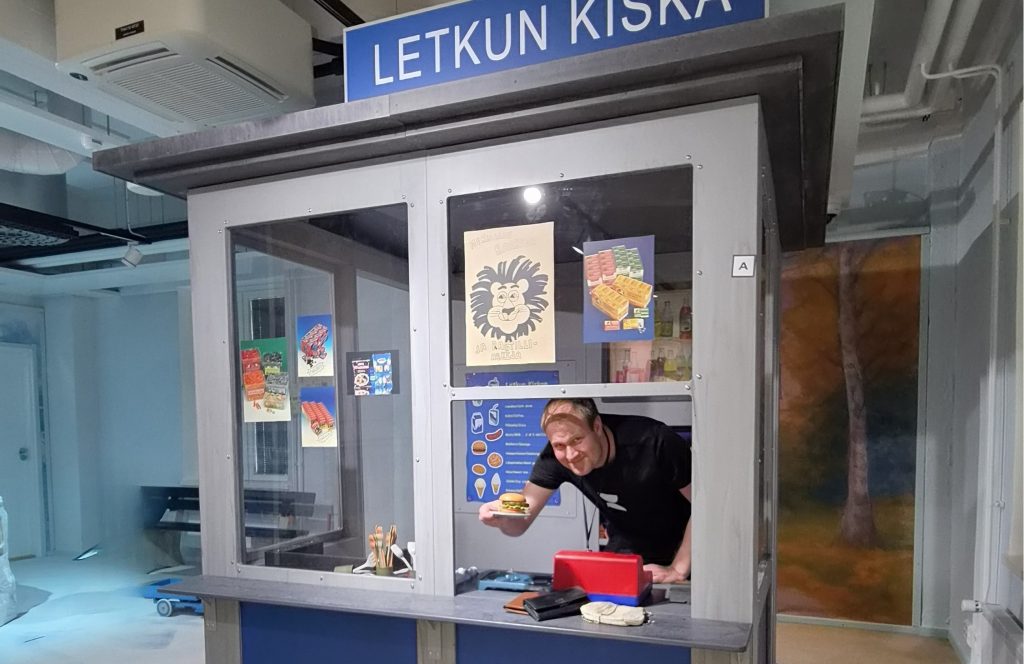 Henkilö tarjoaa leikkihampurilaista kioskin luukusta kurkottaen.